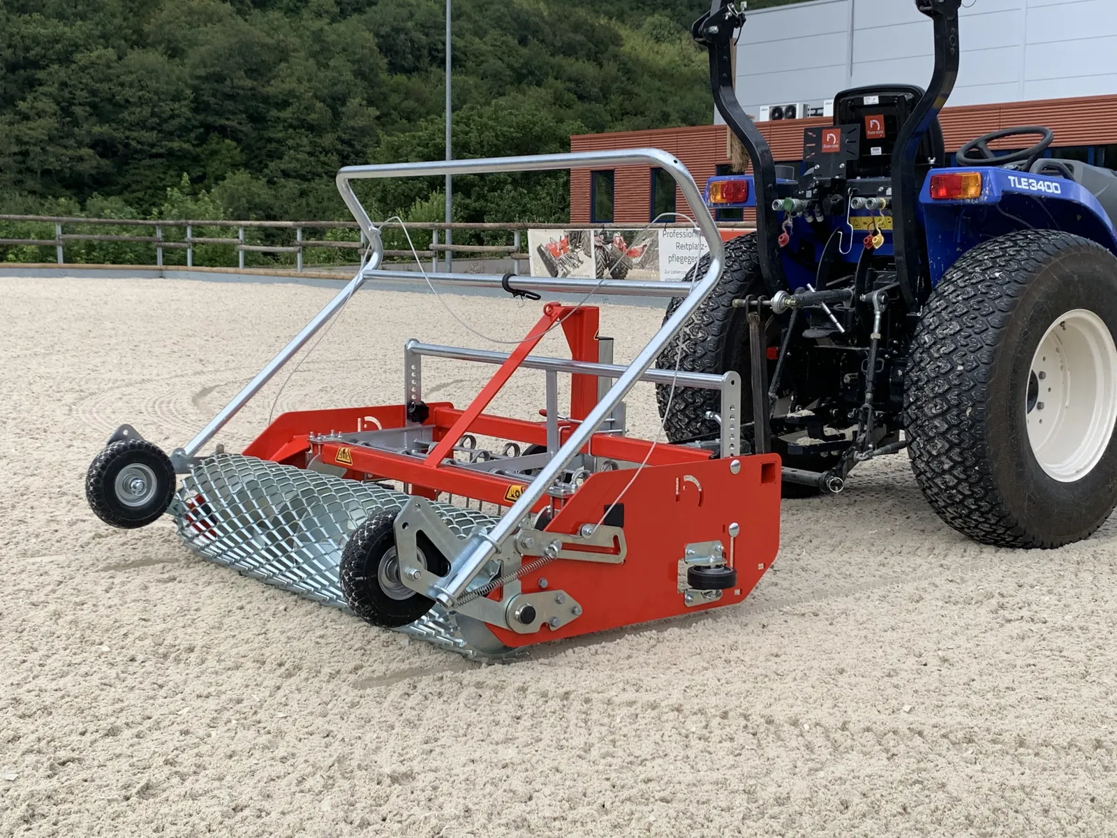 Floor-Care AHK 145/200: rode vlakker met gaasrol en duwbeugel achter een blauwe compacte trekker in een zandbak